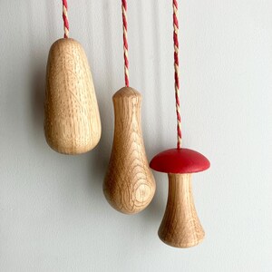 May include: Three wooden light pulls suspended by red and white braided cords. Two are teardrop-shaped, and one is a mushroom shape with a red cap. The pulls are made of light-coloured wood with visible grain.