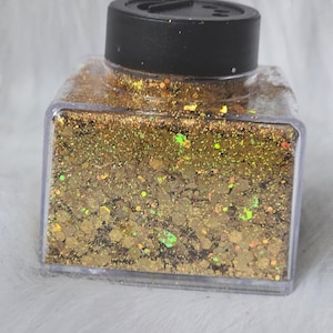 May include: A clear, square container filled with gold glitter. The glitter has a mix of shapes and sizes, with hints of green and orange. The container has a black lid. The glitter is likely used for crafts or decorative purposes.
