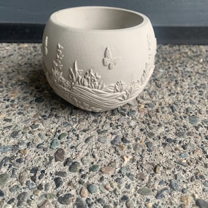 以下が含まれることがあります： 蝶と花の要素が詳細にレリーフされた、丸いオフホワイトのプランター。 プランターは滑らかで未釉薬仕上げで、小さな植物や多肉植物に適しています。 デザインはプランターの外側を包み込んでいます。