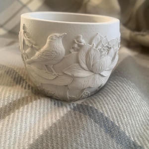 Könnte beinhalten: Kleiner, weißer, zylindrischer Pflanztopf mit detailliertem Reliefdesign. Das Relief zeigt einen Vogel, der auf einem Ast neben einer großen Blume sitzt. Der Topf besteht aus einem matten, festen Material.