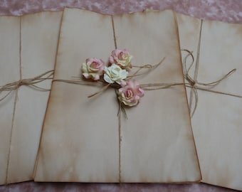 Pode incluir: Uma pilha de três folhas de papel de estilo vintage amarradas com barbante e decoradas com cinco pequenas rosas de papel em tons de rosa e branco.