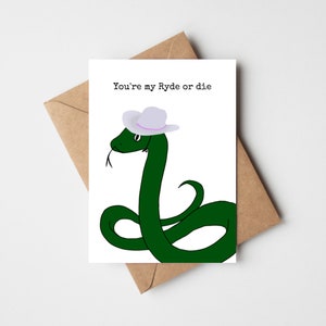 Puede incluir: Tarjeta de felicitación con una serpiente verde de dibujos animados que lleva un sombrero de vaquero lila claro. La tarjeta dice "You're my Ryde or die" en texto negro. La tarjeta está colocada en un sobre marrón.