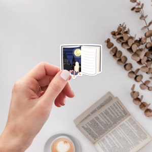 May include: A sticker of an open book with a night scene inside. The scene shows a city skyline with a full moon and a person sitting on a ledge looking out at the scene.