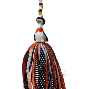 May include: A hanging ornament featuring a small penguin figurine wearing a black hat and orange bowtie, suspended by an orange ribbon. Above the penguin are black, white, and orange beads. Below the penguin is a tassel made of various ribbons.