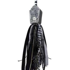 May include: A hanging ornament featuring a grey tombstone-shaped charm with the text "HERE LIES PEPPER" and a skull graphic. The charm is attached to a black ribbon and a cluster of black, white, and grey fabric ribbons, including gingham and metallic strands.