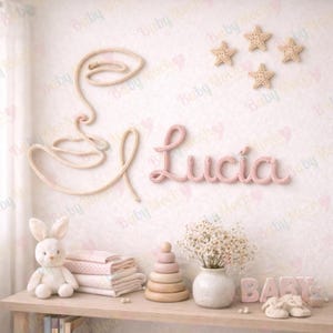 Puede incluir: Decoración de pared para guardería con un contorno de cara tejido, el nombre "Lucia" en hilo rosa y decoraciones en forma de estrella. Una estantería debajo muestra un conejito de peluche, mantas apiladas, un juguete apilable de madera y un jarrón con flores.