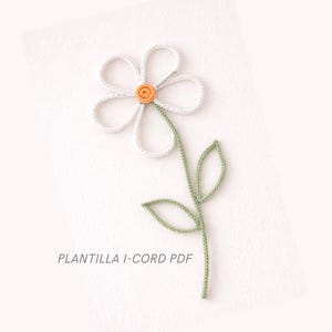 Op de afbeelding: Een gehaakte bloem met witte bloemblaadjes, een oranje centrum en een groene stengel en bladeren. De tekst "PLANTILLA I-CORD PDF" staat onderaan. De bloem is een handgemaakt knutselartikel.