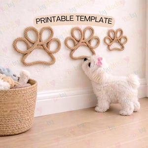 Puede incluir: Tres decoraciones de huellas de pata de cuerda marrón están montadas en una pared blanca, con un perro blanco pequeño con un lazo rosa mirándolas. Una cesta tejida llena de animales de peluche está en el suelo. El texto "PRINTABLE TEMPLATE" está arriba.