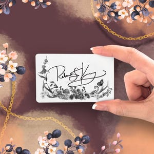 Puede incluir: Una tarjeta de visita blanca con un diseño floral y la firma "Rebecca S. King" en negro. La tarjeta es sostenida por una mano sobre un fondo con detalles florales y una cadena dorada.