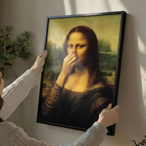 Könnte beinhalten: Gerahmter Druck der Mona Lisa, humorvoll verändert, um sie mit zugehaltener Nase zu zeigen. Das Kunstwerk befindet sich in einem schwarzen Rahmen, der von einer Person gehalten wird. Der Hintergrund zeigt eine Wand und einige Pflanzen.