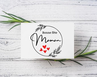 May include: A rectangular white greeting card featuring a black circular wreath design with leafy accents. The text reads Bonne fête Maman in black script and print, accompanied by four small red hearts centered within the circular frame.