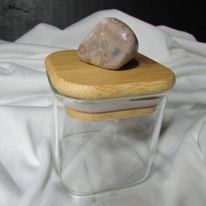 May include: A square glass jar with a bamboo lid and a decorative stone on top. The jar is clear and the lid is a light brown color. The stone is a mottled pink and brown color.