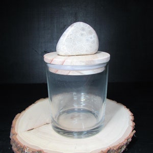 May include: A clear glass jar with a wooden lid and a decorative stone on top. The jar sits on a round wooden slice with bark edges. The background is a dark, solid color.