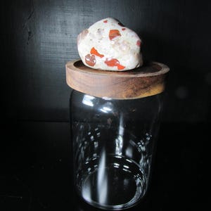 May include: A clear glass jar with a wooden lid. The lid has a red and white speckled stone on top.