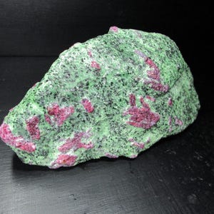 May include: A green and pink rough gemstone with red ruby inclusions. The stone has a rough, irregular shape and is a natural specimen.