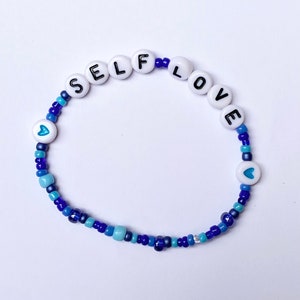 May include: A blue and white beaded bracelet with the words "SELF LOVE" spelled out in white beads with black lettering. The bracelet also features two small blue heart beads.