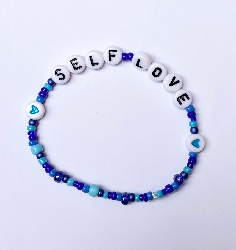 May include: A blue and white beaded bracelet with the words "SELF LOVE" spelled out in white beads with black lettering. The bracelet also features two small blue heart beads.