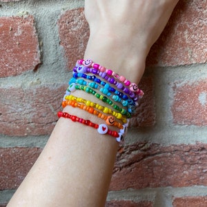 May include: A stack of colourful beaded bracelets with smiley face and heart charms. The bracelets are layered on a person's wrist, creating a rainbow effect.