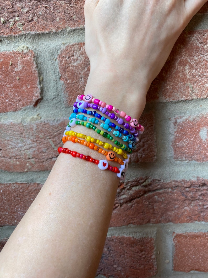 May include: A stack of colourful beaded bracelets with smiley face and heart charms. The bracelets are layered on a person's wrist, creating a rainbow effect.