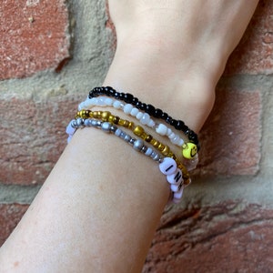 May include: A stack of beaded bracelets on a wrist. The bracelets are black, white, gold, and silver. One bracelet has a smiley face charm and another has the word "INH" spelled out in white beads.