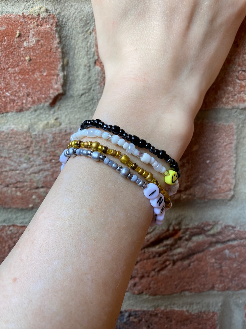 May include: A stack of beaded bracelets on a wrist. The bracelets are black, white, gold, and silver. One bracelet has a smiley face charm and another has the word "INH" spelled out in white beads.