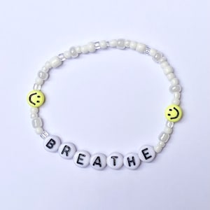 May include: White beaded bracelet with the word "BREATHE" spelled out in black letters on white beads. The bracelet also has two yellow smiley face beads.