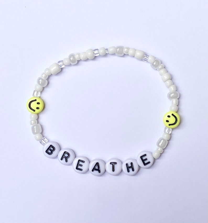 May include: White beaded bracelet with the word "BREATHE" spelled out in black letters on white beads. The bracelet also has two yellow smiley face beads.