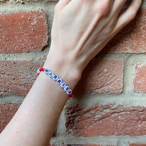 May include: A red and white beaded bracelet with the word "WARRIOR" spelled out in black letters.