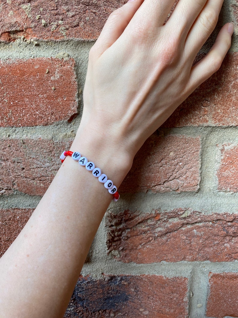 May include: A red and white beaded bracelet with the word "WARRIOR" spelled out in black letters.