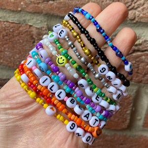May include: A stack of colourful beaded bracelets with letter and smiley face charms. The bracelets spell out "I'll Be At 90" and "Green" with a smiley face charm on each bracelet.