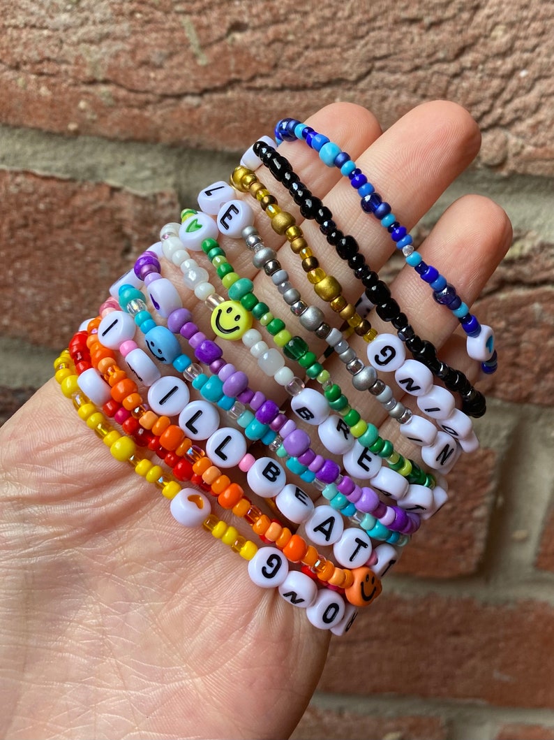 May include: A stack of colourful beaded bracelets with letter and smiley face charms. The bracelets spell out "I'll Be At 90" and "Green" with a smiley face charm on each bracelet.