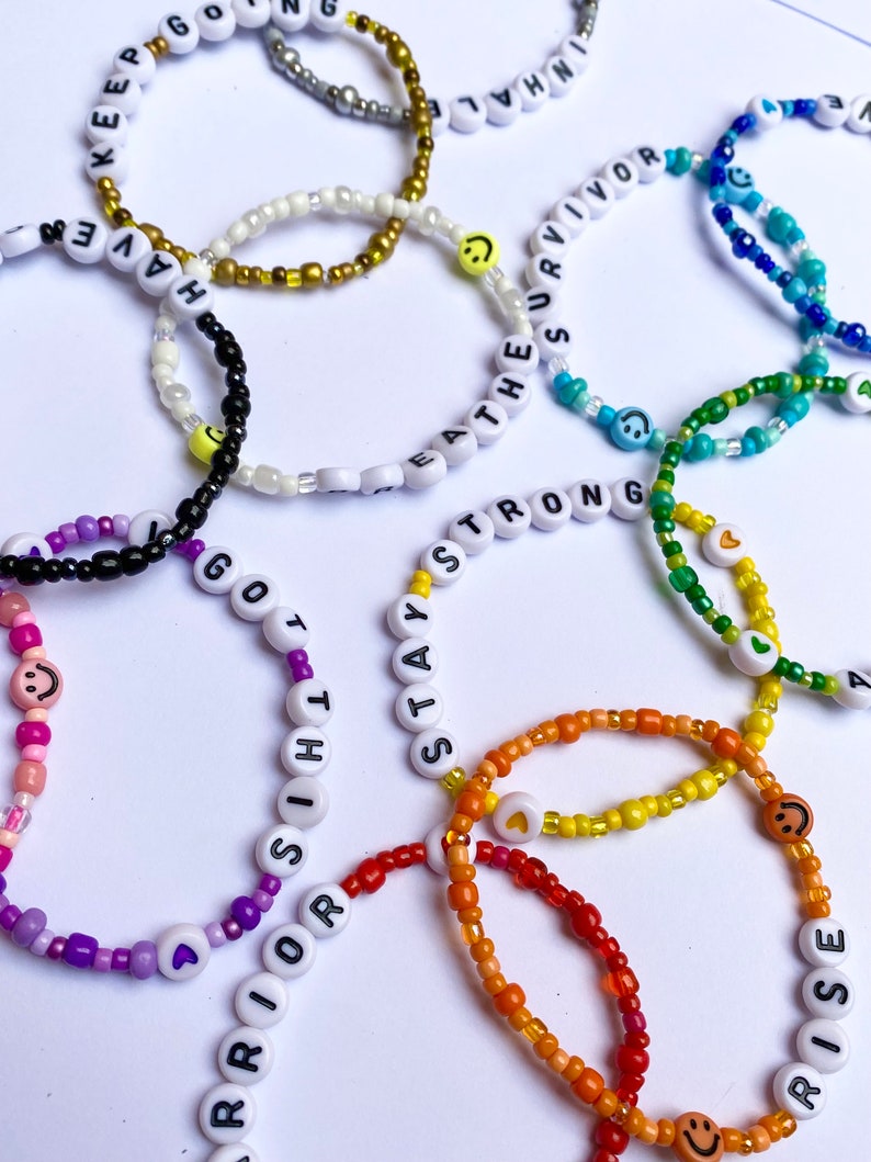 May include: A collection of colourful beaded bracelets with positive messages, including "Keep Going", "Have Faith", "Stay Strong", "Warrior", "Rise", "Breathe", "Eat The Survivor", and "Go With It".
