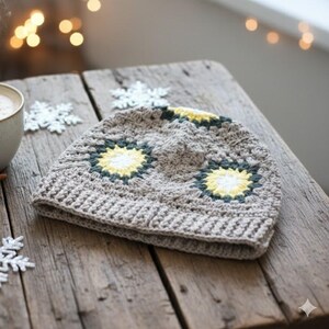 Può includere: Una scena accogliente con un berretto all'uncinetto grigio con dettagli floreali gialli e verdi, una tazza di caffè fumante, bastoncini di cannella e fiocchi di neve decorativi su una superficie di legno rustico. Il berretto è fatto a mano.