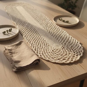 Op de afbeelding: Een beige gehaakte tafelloper met een centraal effen paneel en opengewerkte randen, weergegeven op een houten tafel. Twee bijpassende borden met takjes en een gevouwen linnen servet maken de rustieke tafelsetting compleet.