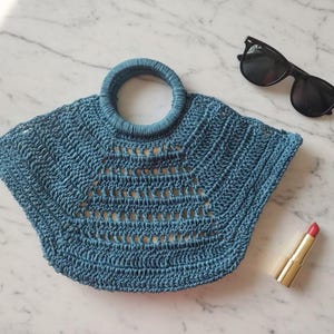 May include: A blue crocheted handbag with a circular handle and an open-work pattern, accompanied by black sunglasses and a gold-cased lipstick. The bag features a wide, fan-like shape with a textured stitch design throughout the body.