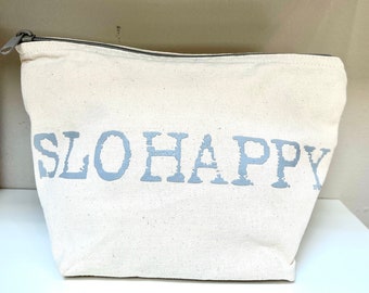 Könnte beinhalten: Eine weiße Canvas-Reißverschlusstasche mit einem grauen Aufdruck, der "SLO HAPPY" lautet.