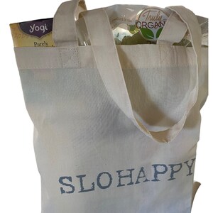K&ouml;nnte beinhalten: Eine wei&szlig;e Canvas-Tragetasche mit dem Text "SLOHAPPY" in dunkelgrau gedruckt. Die Tasche hat zwei Henkel und ist gef&uuml;llt mit Lebensmitteln, darunter eine Schachtel Yogi-Tee und eine T&uuml;te Bio-Produkte.