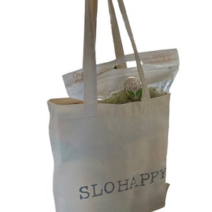 K&ouml;nnte beinhalten: Eine wei&szlig;e Canvas-Tragetasche mit dem Aufdruck "SLOHAPPY" auf der Vorderseite. Die Tasche ist mit einer T&uuml;te gr&uuml;ner kernloser Trauben gef&uuml;llt.