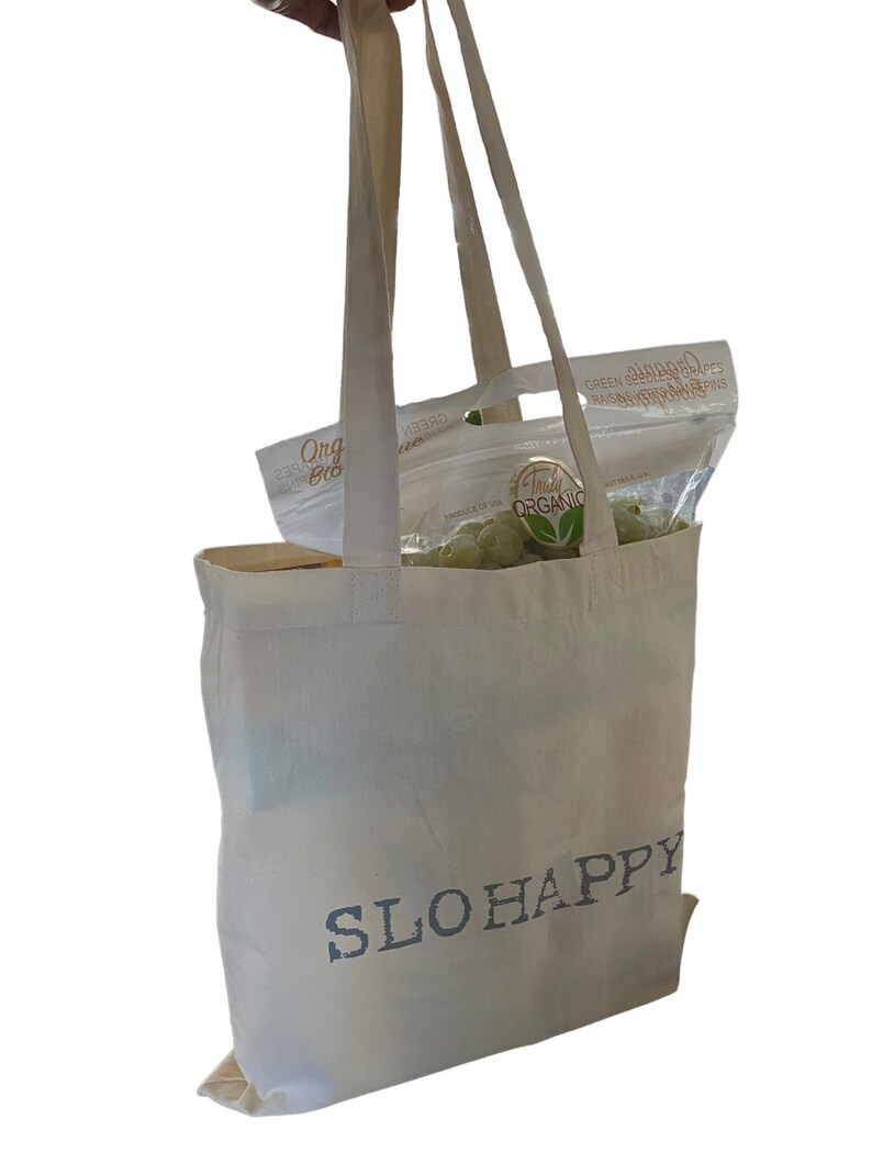 K&ouml;nnte beinhalten: Eine wei&szlig;e Canvas-Tragetasche mit dem Aufdruck "SLOHAPPY" auf der Vorderseite. Die Tasche ist mit einer T&uuml;te gr&uuml;ner kernloser Trauben gef&uuml;llt.