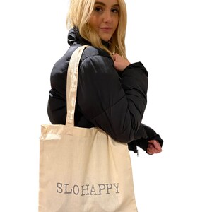 K&ouml;nnte beinhalten: Eine beige Canvas-Tragetasche mit dem Aufdruck "SLOHAPPY" in Grau auf der Vorderseite.