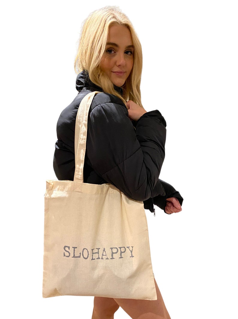 K&ouml;nnte beinhalten: Eine beige Canvas-Tragetasche mit dem Aufdruck "SLOHAPPY" in Grau auf der Vorderseite.