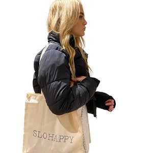 K&ouml;nnte beinhalten: Eine Person h&auml;lt eine beige Tragetasche mit dem Text "SLOHAPPY" in grau bedruckt. Die Tasche hat schwarze Henkel.