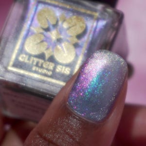 May include: A clear glass bottle of nail polish with a silver iridescent finish. The bottle has a black cap and a gold label that reads "Glitter Sis Studio". A fingernail is painted with the polish, showing the iridescent finish.