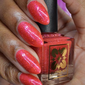 May include: A square glass bottle of bright coral nail polish with a black cap, featuring a gold label with a four-leaf clover design and the text GLITTER SIS STUDIO. The polish has a high-shine finish with fine, reflective glitter particles.