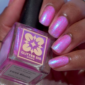 May include: A bottle of pink nail polish with a holographic finish. The bottle is labeled "Glitter Sis Studio Nail Lacquer First Blush 15ml/0.5 fl oz".