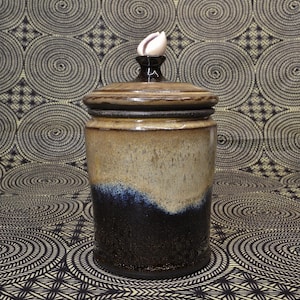 May include: A ceramic jar with a lid, featuring a gradient of colors from tan to dark blue. The jar is topped with a dark brown finial and a white seashell. The jar has a cylindrical shape and a textured surface.