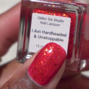 May include: A square glass bottle of Glitter Sis Studio nail lacquer with a white label reading I Am Hardheaded & Unstoppable and 15 ml. The polish is a vibrant red with reflective gold and orange glitter particles throughout the liquid.