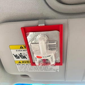 May include: A red plastic holder clipped to a car visor, containing a nasal spray and other medical supplies. The nasal spray is labeled with "4mg" and "NASAL SPRAY". The visor has warning labels.