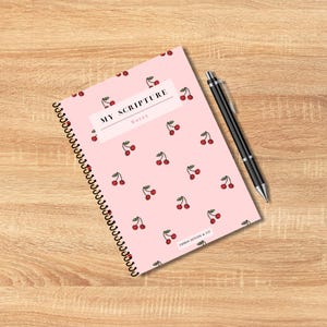May include: A spiral-bound notebook with a pink cover adorned with cherry illustrations. The text "MY SCRIPTURE Notes" is displayed on the cover. A black pen rests beside the notebook on a wooden surface. The notebook is from Ember Design & Co.