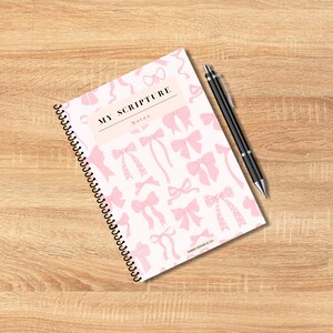 May include: A spiral-bound notebook with a cover featuring a pattern of pink bows on a white background. The text "MY SCRIPTURE Notes" is printed on the cover. A black pen rests on the wooden surface next to the notebook.